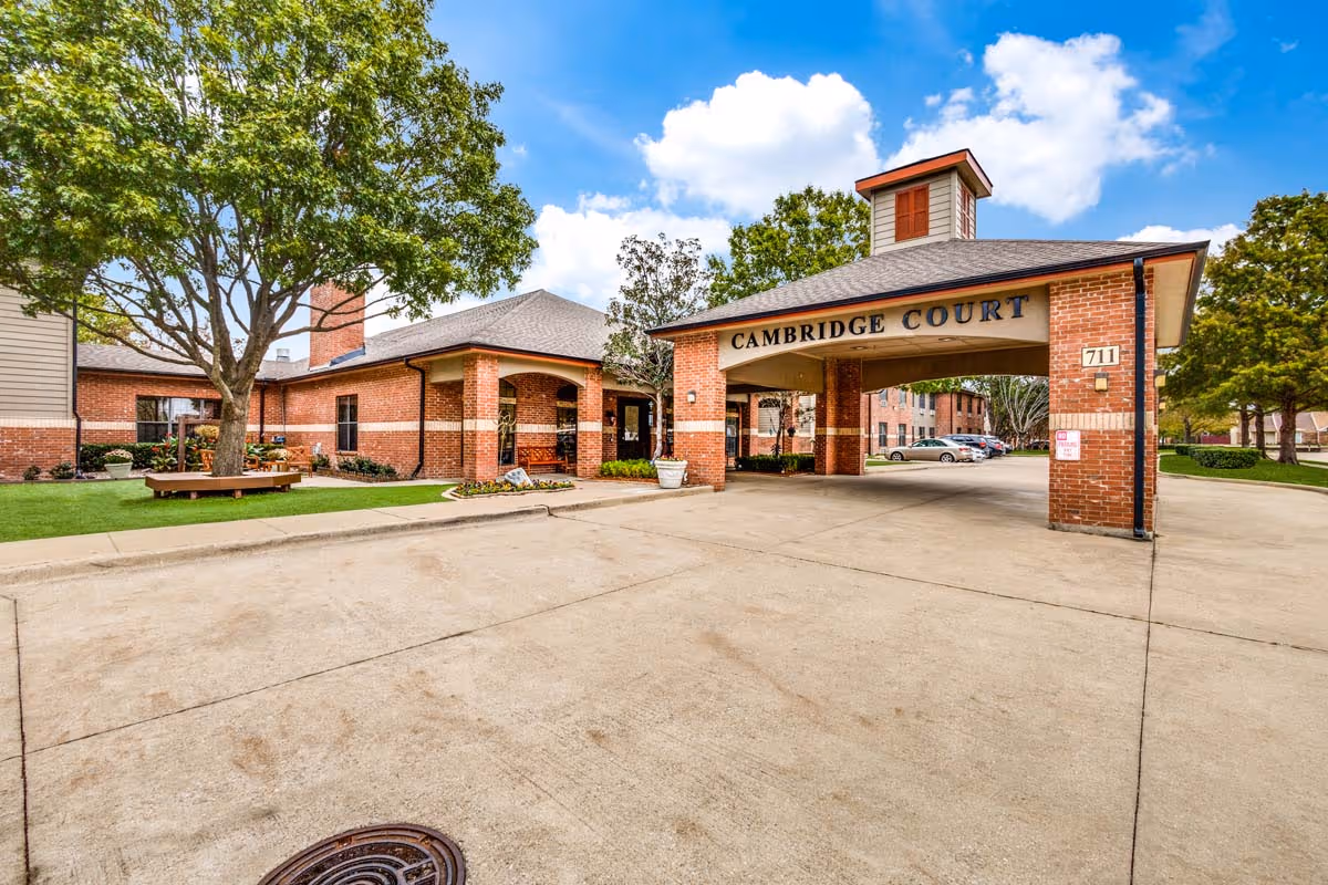 Exterior view of Cambridge Court Senior Living facility showing a brick building with an arched entrance labeled 'Cambridge Court'. There is a large tree with a circular bench around it, a paved driveway, parked cars, and a bright blue sky with some clouds.