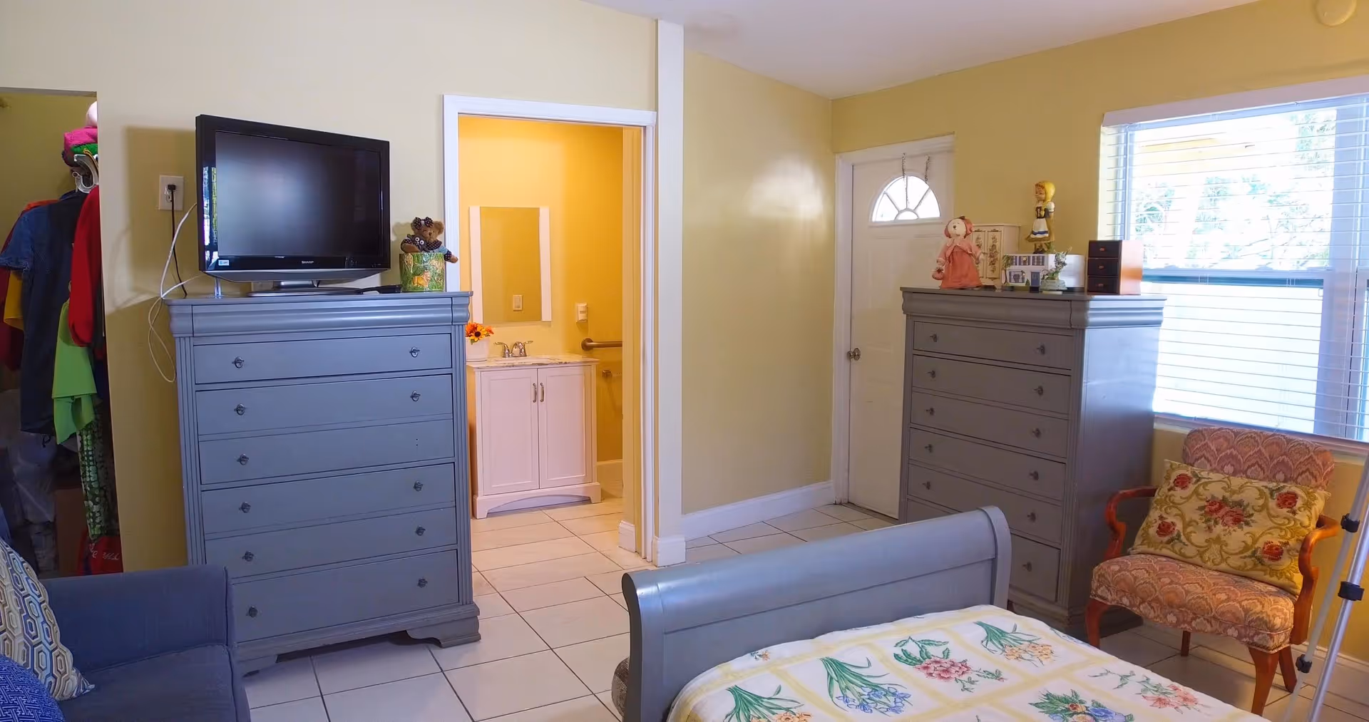 Bedroom with two gray dressers, a bed, TV, armchair, and an open doorway leading to a bathroom.