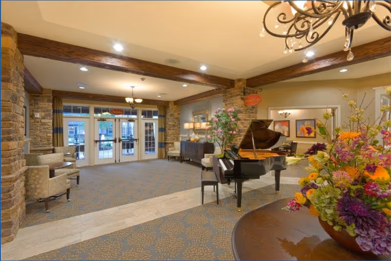 A spacious and warmly decorated interior common area featuring a grand piano with a bench, floral arrangements on a round wooden table, comfortable armchairs, stone pillars, and large glass doors leading outside. The ceiling has wooden beams and a chandelier, creating an inviting atmosphere.