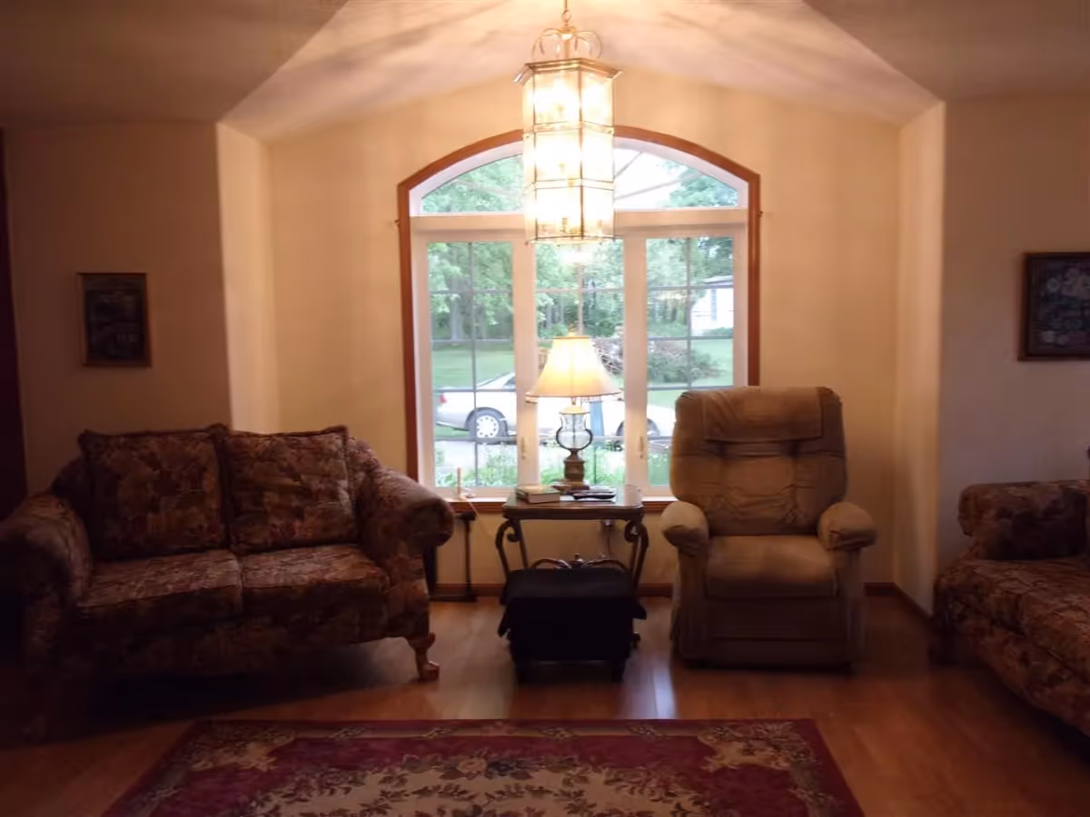 A cozy living room with two floral-patterned sofas on either side and a beige recliner chair in the center. Between the recliner and one sofa is a small wooden side table with a lamp, books, and other items. Behind the furniture is a large window with an arched top, showing a view of a green yard and a parked car outside. A hanging light fixture is visible from the ceiling, and a patterned rug covers part of the wooden floor.