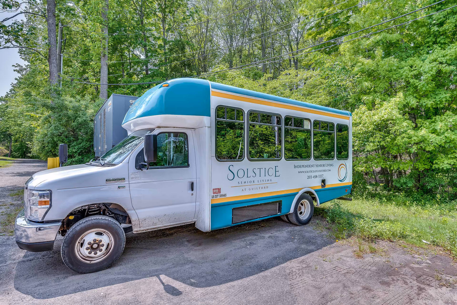 A white and teal shuttle bus parked on a gravel area surrounded by green trees. The bus has the text 'Solstice Senior Living at Guilford' and contact information on its side, indicating it is used for independent senior living transportation.