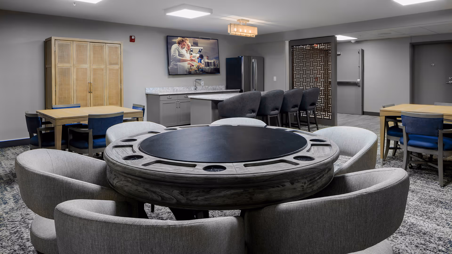 A modern common area with a large round table surrounded by six cushioned chairs in the foreground. In the background, there are two wooden tables with blue chairs, a kitchen island with four bar stools, a refrigerator, and a wall-mounted TV displaying a cooking scene. The room has gray walls, carpeted floors, and a decorative wooden partition near a hallway.