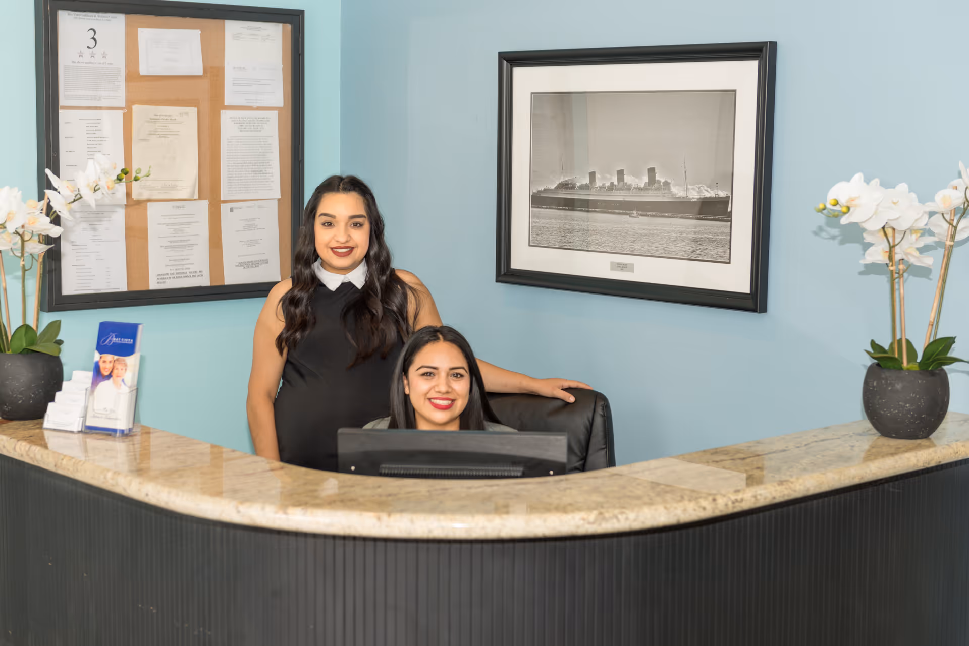 Two women smiling behind a reception desk in a healthcare facility. The desk has a granite countertop with potted white orchids on each side. Behind them is a bulletin board with papers pinned and a framed black-and-white photograph of a ship on a light blue wall.