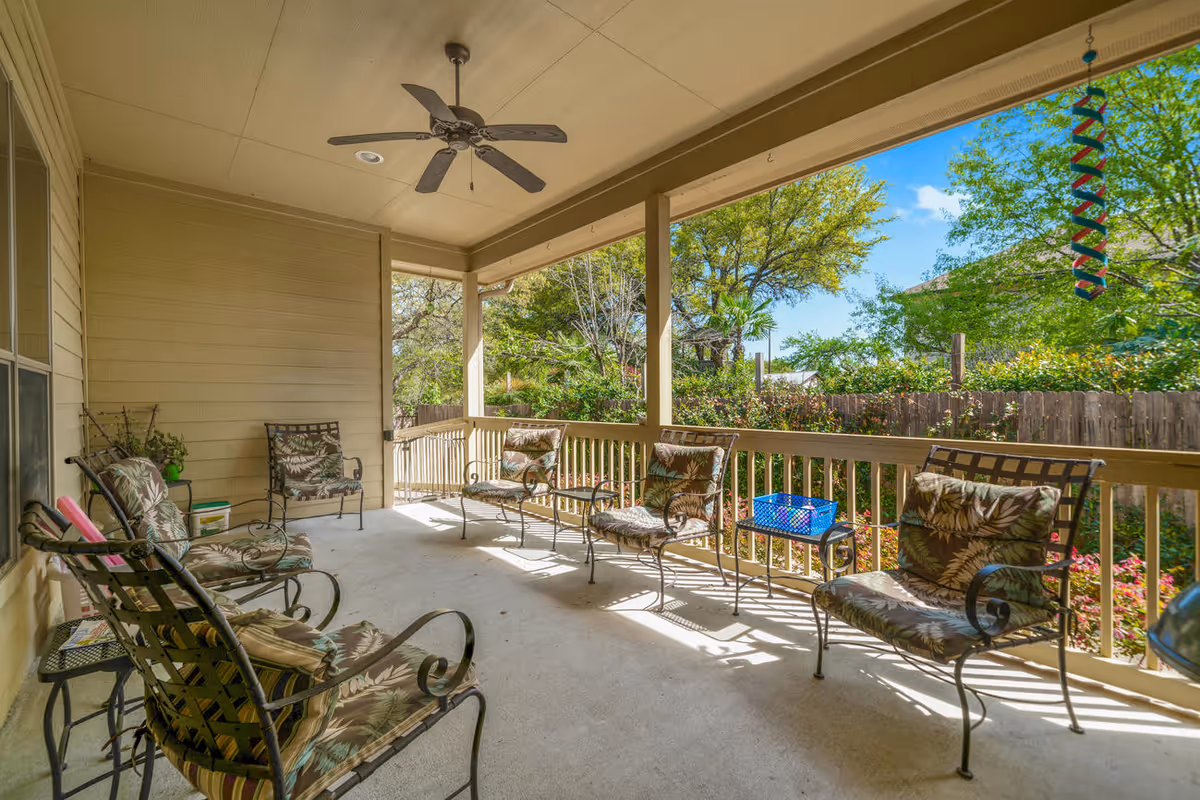 Covered outdoor patio area with several metal chairs featuring floral cushions arranged along the railing and wall. A ceiling fan is mounted on the ceiling, and there is a view of trees and greenery beyond the wooden fence surrounding the patio.
