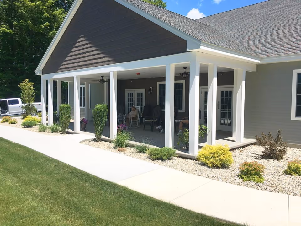 Exterior view of Beaver Lake Lodge Assisted Living showing a covered porch area with white pillars, outdoor seating, and some people sitting. The building has gray siding and a dark shingled roof. There are landscaped bushes and plants along the walkway and a green lawn in the foreground.