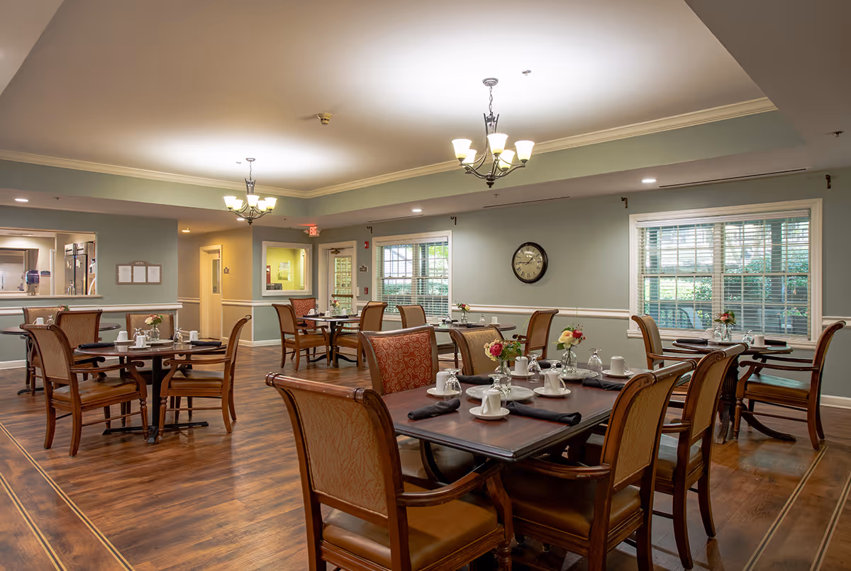 A well-lit dining room in a senior living facility with several wooden tables and chairs arranged neatly. Each table is set with white cups, saucers, black napkins, and small flower vases. The room has large windows with white blinds, light green walls, a clock on the wall, and two chandeliers hanging from the ceiling. The floor is wooden, and there is a serving window to the kitchen area.