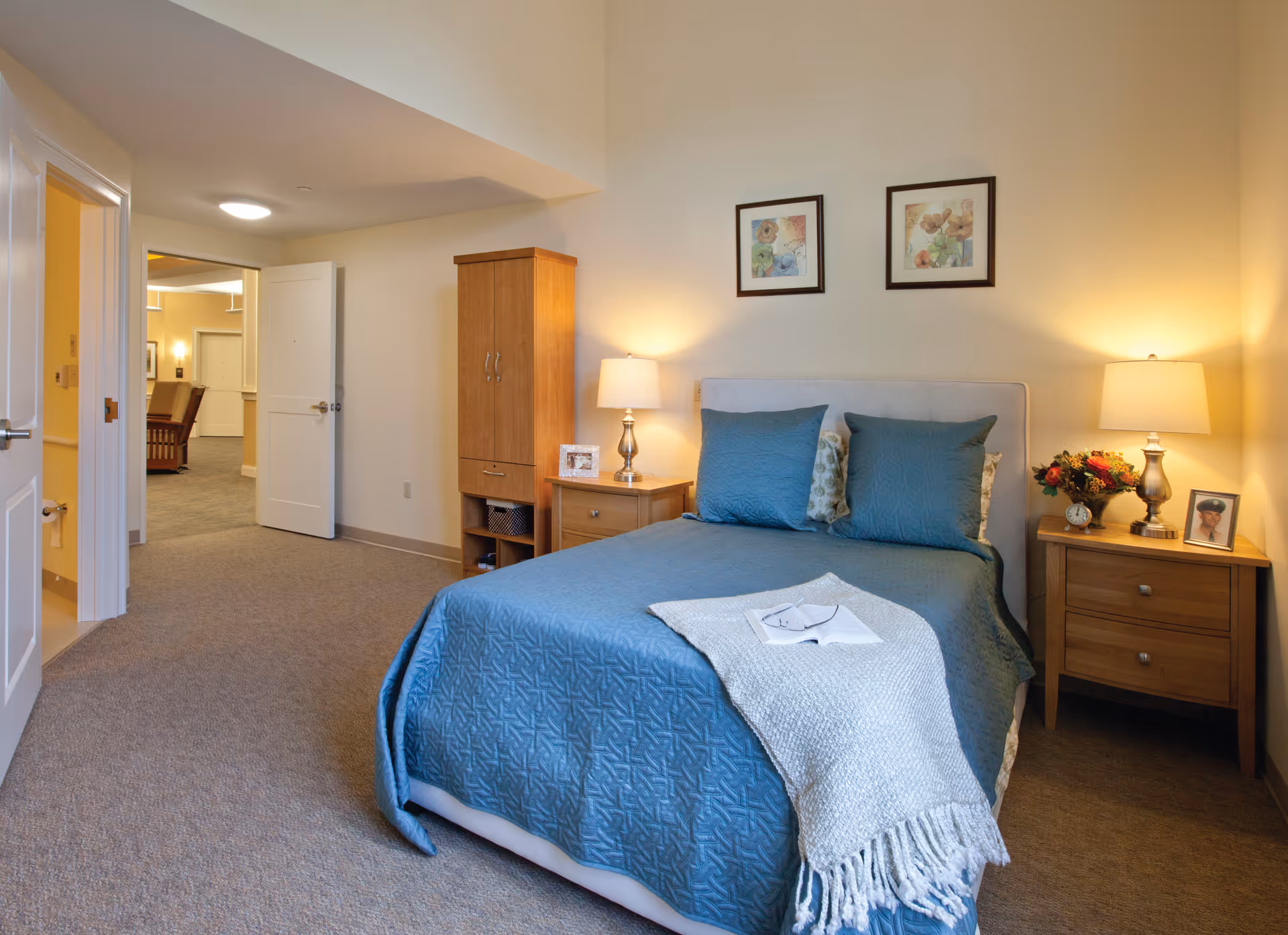 A neatly furnished bedroom with a blue-covered bed, bedside tables and lamps, a wardrobe, and an open door to a hallway.
