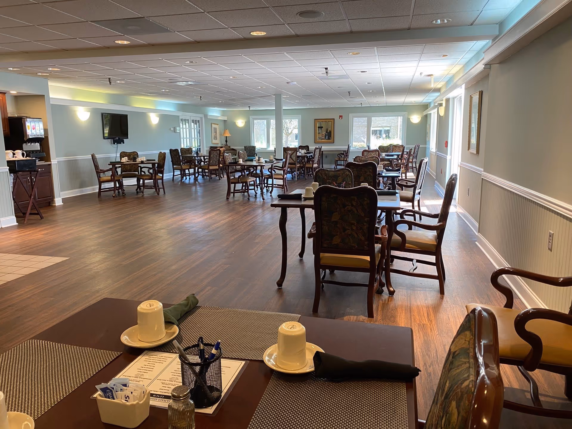 A spacious dining room with multiple wooden tables and chairs arranged neatly. Each table has placemats, cups, and napkins. The room has large windows letting in natural light, light blue walls with white wainscoting, and a flat-screen TV mounted on one wall. There is a beverage station with a drink dispenser on the left side of the room.
