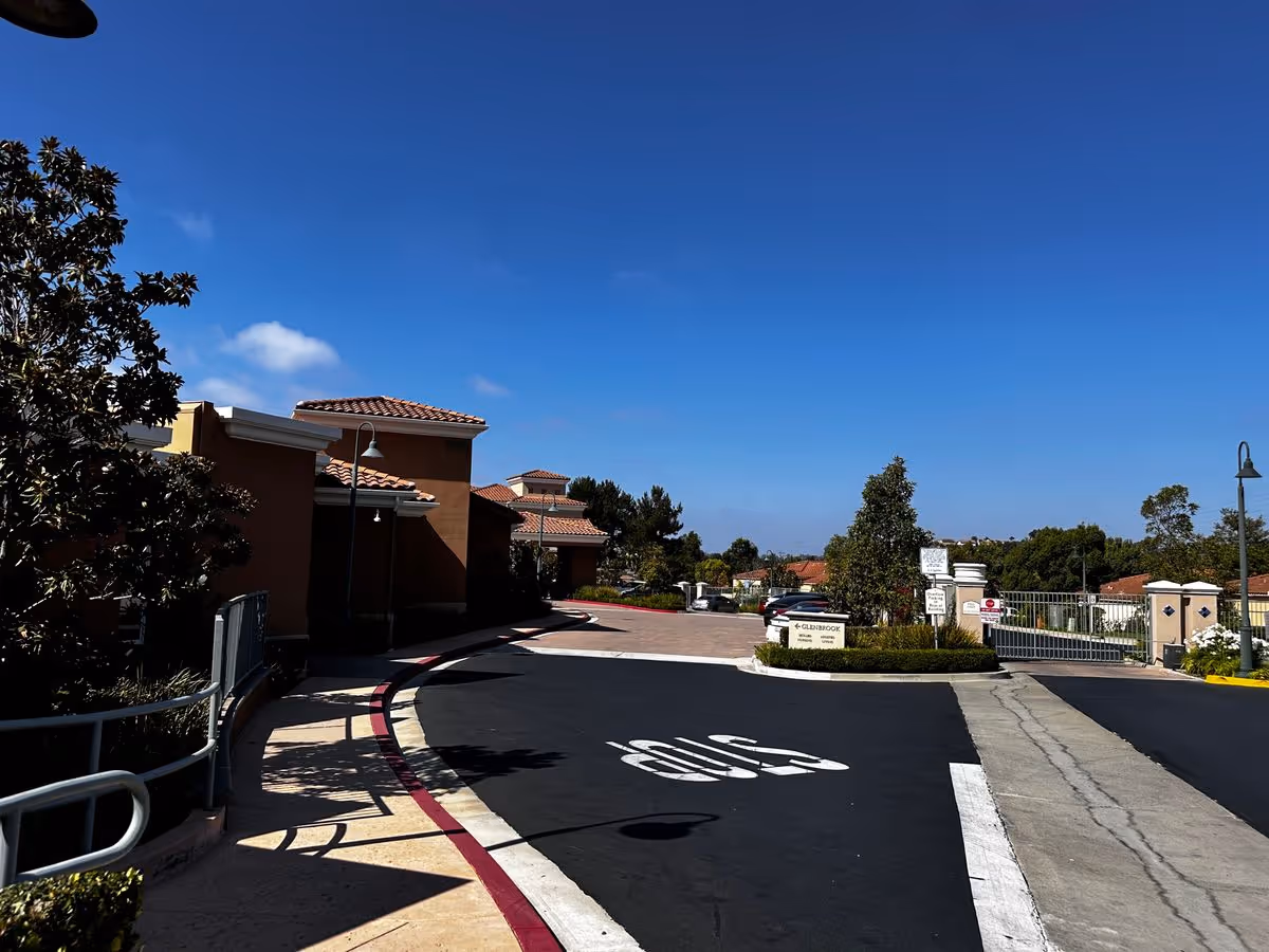 Driveway entrance to GlenBrook Health Center with tiled-roof buildings, gated entrance, landscaping, and a painted STOP on the road under a clear blue sky.