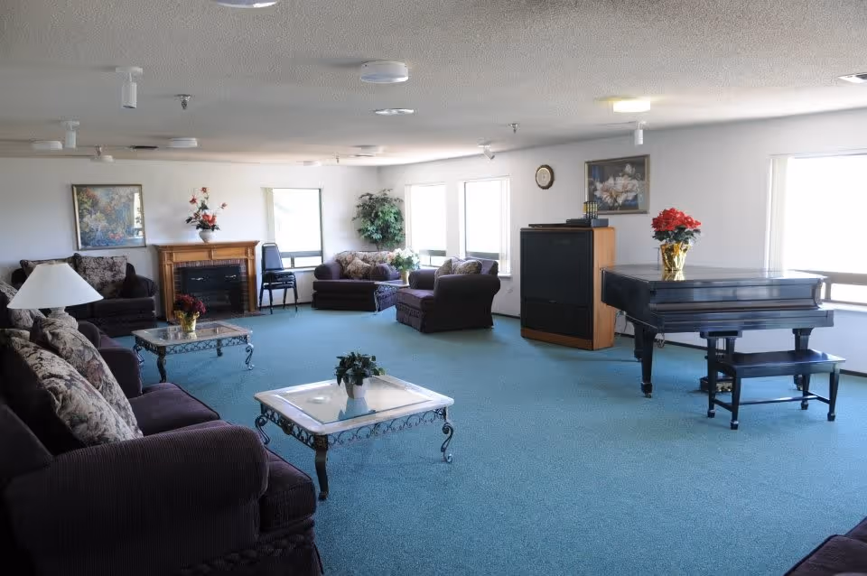 A spacious living room with teal carpet, multiple cushioned armchairs and sofas with floral patterned pillows, two glass-top coffee tables with decorative plants, a black grand piano with a red poinsettia on top, a wooden cabinet with a clock and books, a fireplace with a floral arrangement, and several windows letting in natural light.