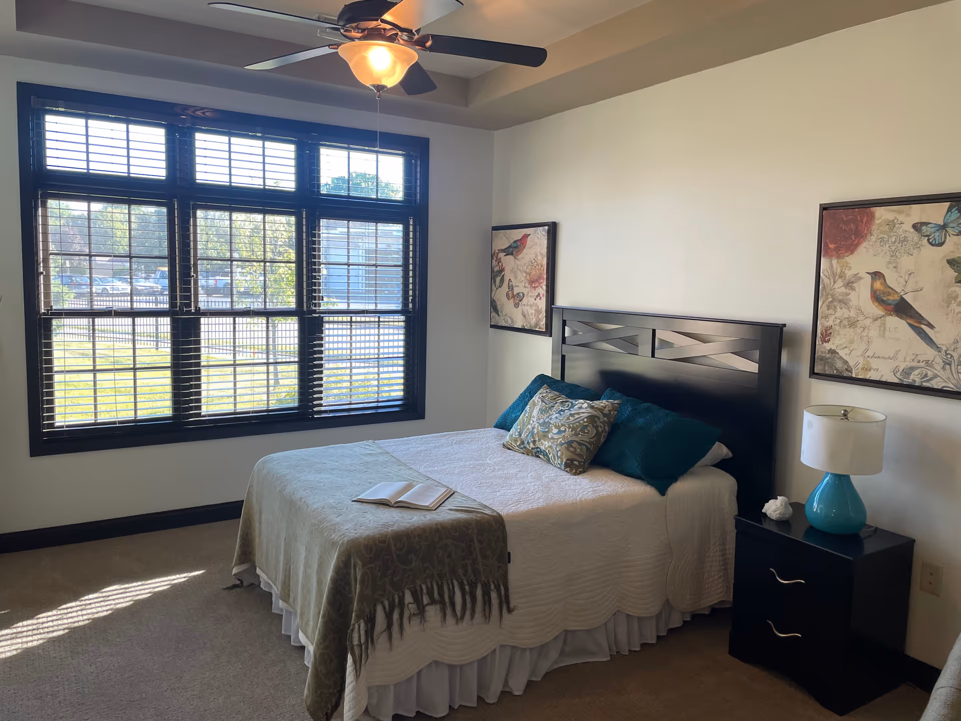 A bedroom with a bed covered in white and green bedding, two teal pillows, and a decorative pillow. There is a nightstand with a blue lamp and a small decorative item. Two framed pictures featuring birds and butterflies hang on the wall above the bed. A large window with black trim and blinds lets in natural light. A ceiling fan with a light fixture is mounted on the ceiling.