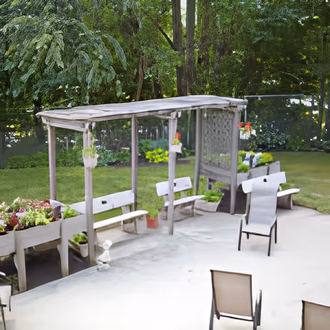 Outdoor patio area with a wooden pergola structure featuring hanging plants and benches underneath. There are raised garden beds with various plants and flowers, surrounded by lush green trees and grass in the background. Two outdoor chairs are positioned on a concrete surface in the foreground.