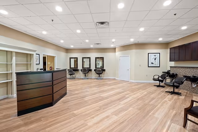 Interior view of a spacious salon area with wooden flooring, a reception desk on the left, three styling stations with mirrors and chairs along the back wall, and three black salon chairs with sinks on the right side. The ceiling has recessed lighting and the walls are painted beige with white trim. There is a framed picture on the right wall and a closed white door in the center background.