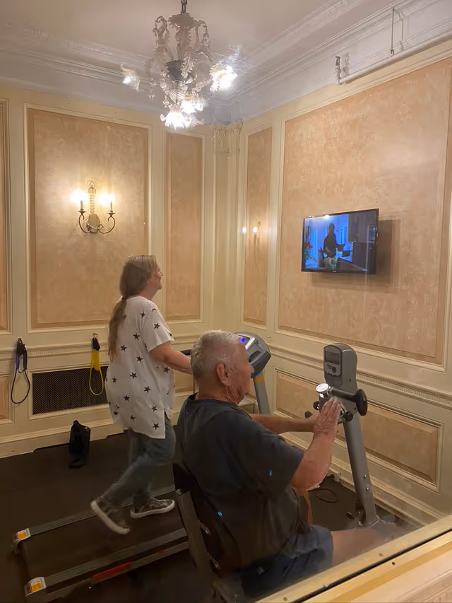 Two seniors exercising on a treadmill and a recumbent bike in an ornate exercise room with a wall-mounted TV.