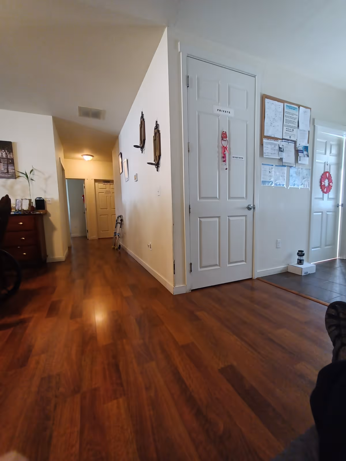 Interior hallway of a senior living facility with wooden flooring, white walls, and several doors. One door is labeled 'PRIVATE' with a 'DO NOT ENTER' sign. There are bulletin boards with papers on the wall near the doors, and some decorative items such as a small plant and wall hangings are visible.