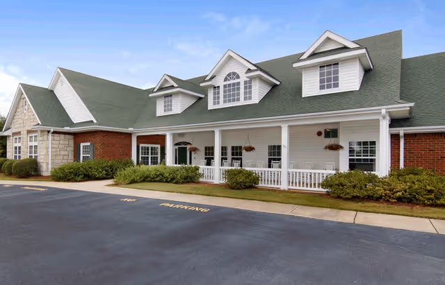 Front exterior of a single-story senior living building with a covered porch, dormer windows, shrubs, and a parking area.