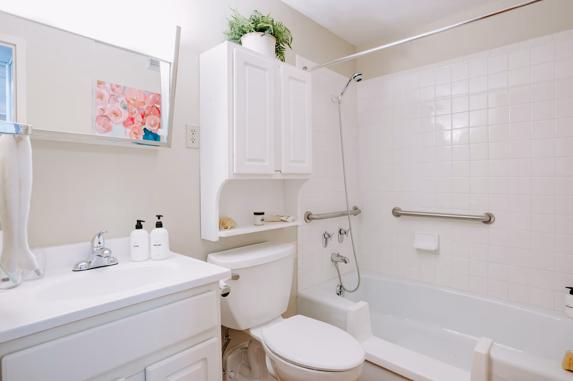 A clean and bright bathroom featuring a white sink with two soap dispensers, a toilet, and a bathtub with a handheld showerhead. The bathtub has safety grab bars installed on the tiled wall. Above the toilet is a white cabinet with a potted plant on top. A towel hangs on a rack next to the sink, and a mirror is mounted on the wall above the sink reflecting a floral painting.