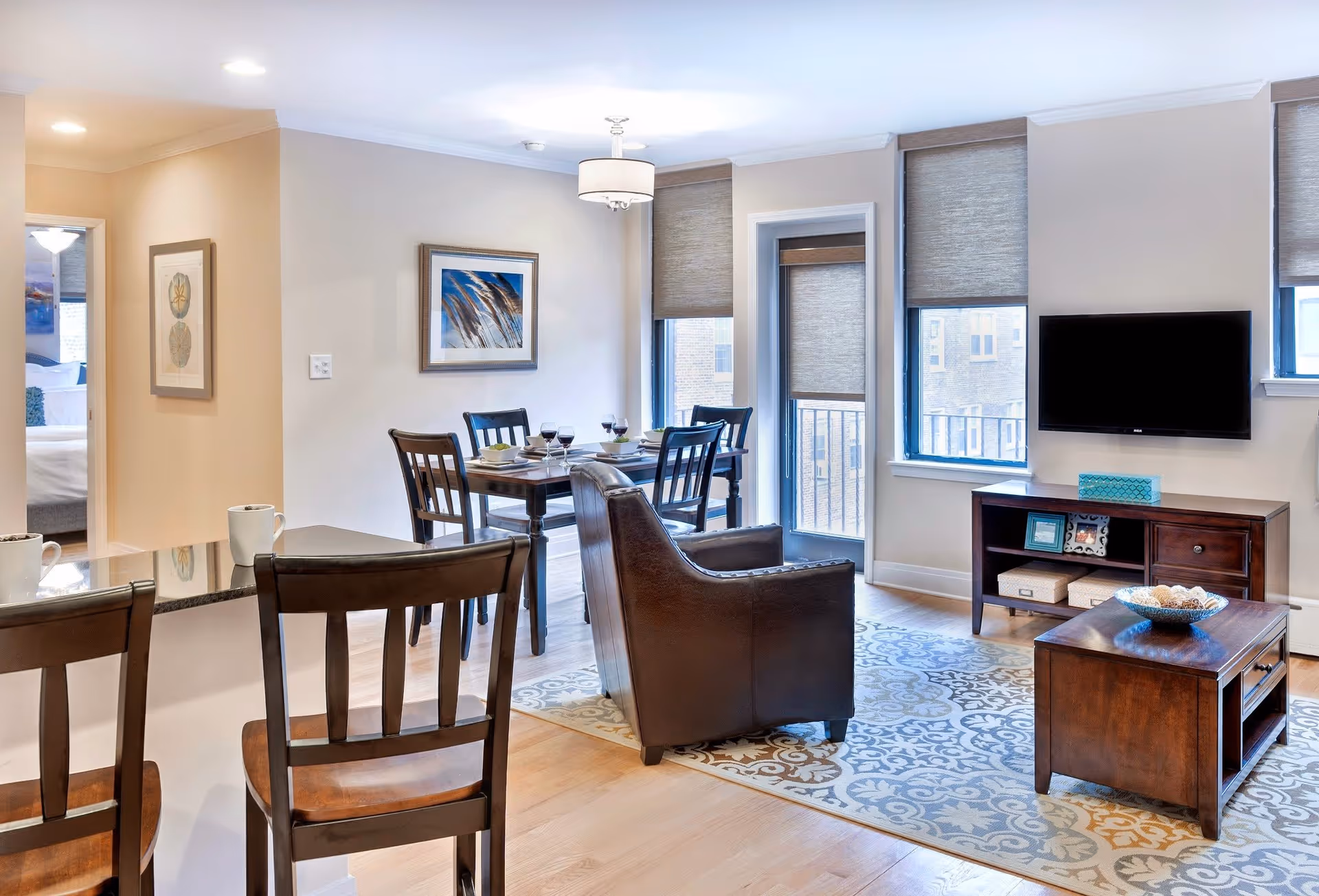 A bright and modern living and dining area in a senior living facility. The space features a wooden dining table set with four chairs and place settings, a dark leather armchair, a wooden coffee table with a decorative bowl, and a TV mounted on the wall above a wooden console. Large windows with beige roller shades allow natural light to fill the room. The floor is light wood with a patterned area rug, and there is a glimpse of a bedroom through an open doorway.