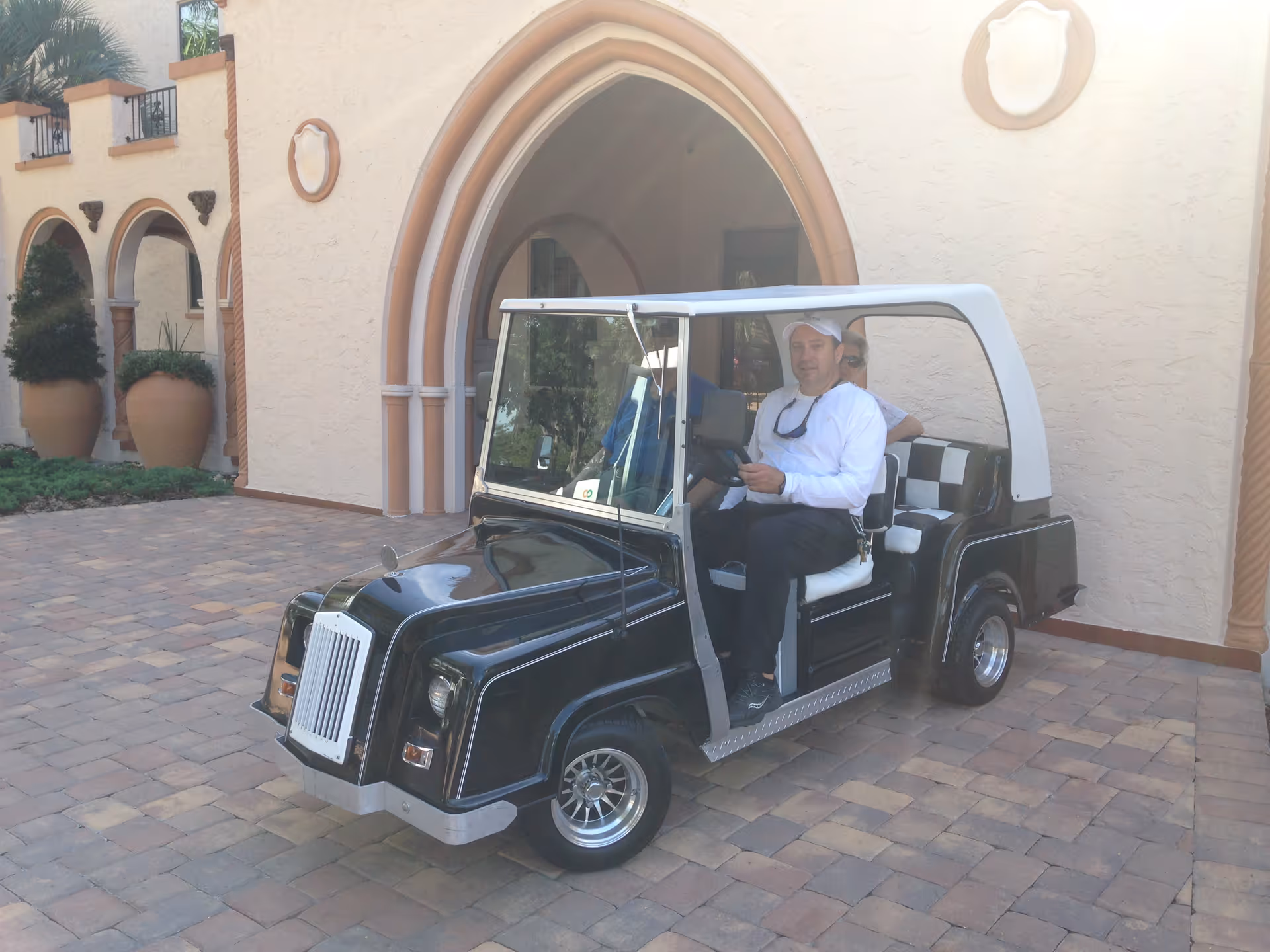 Two men sitting in a black and white golf cart parked on a paved area in front of a building with arched doorways and large potted plants.