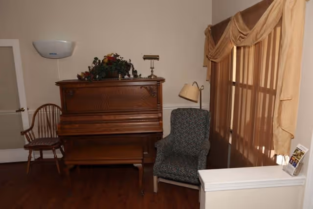 A cozy interior corner featuring a wooden upright piano with a floral arrangement on top, a wooden chair to the left, a patterned armchair to the right, a floor lamp behind the armchair, and a window with beige curtains allowing soft light into the room.