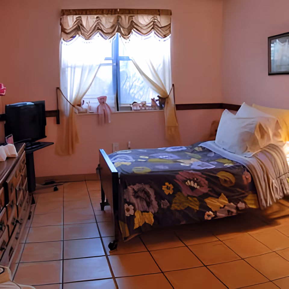A cozy bedroom with a single bed covered in a floral blanket, white pillows, and a striped throw. The room has tiled flooring, a window with sheer curtains tied back, a wooden dresser with a TV on top, and soft pink walls with a framed picture.