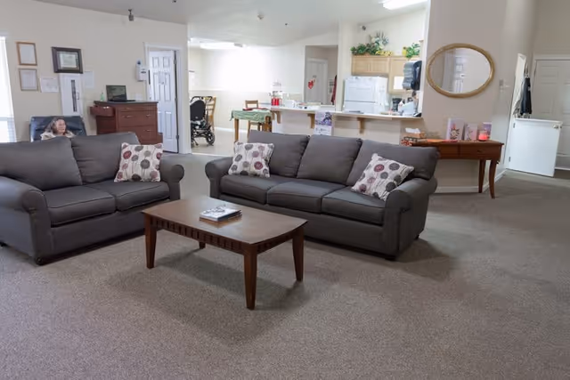 A communal living area with two gray sofas, a wooden coffee table, and a kitchenette and dining area in the background.