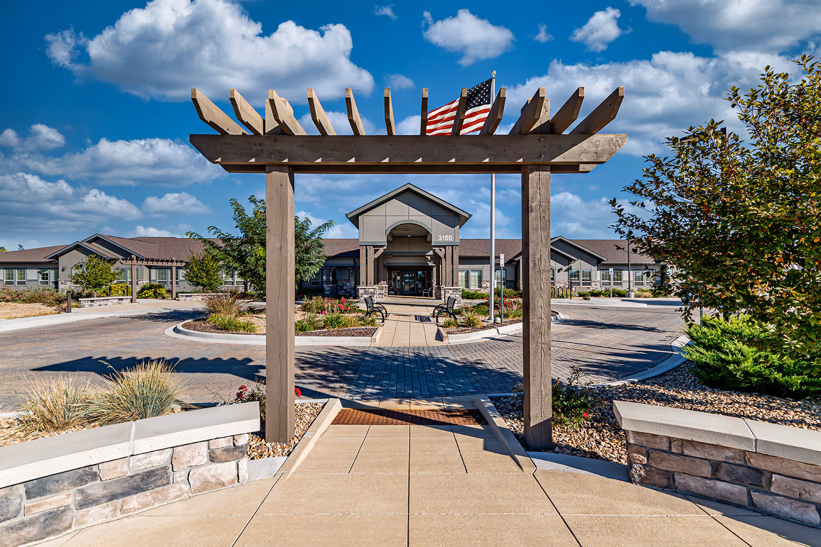 View of the entrance to a senior living facility named Aspens at Fort Collins Memory Care, featuring a wooden pergola structure in the foreground, a paved walkway leading to the building entrance, landscaped gardens with bushes and flowers, an American flag on a flagpole, and a partly cloudy blue sky.