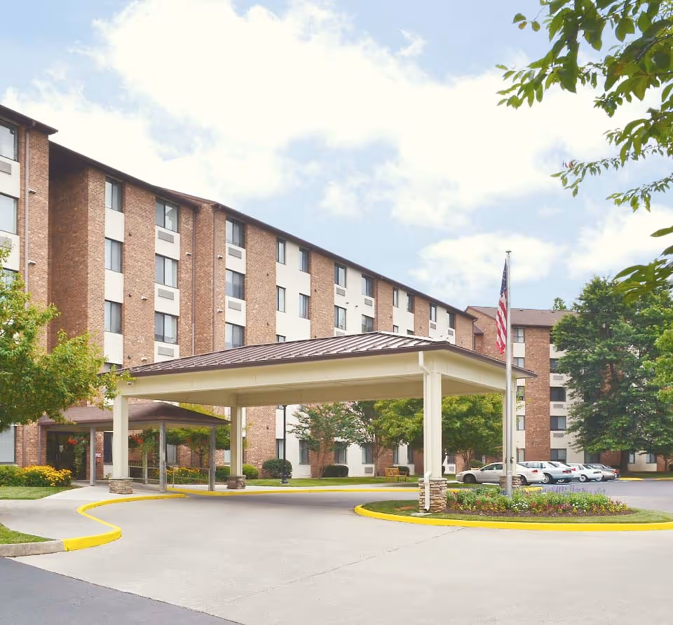 Exterior view of Edinburgh Square senior living facility showing a multi-story brick building with white window panels, a covered driveway entrance, landscaped greenery, and an American flag on a flagpole.