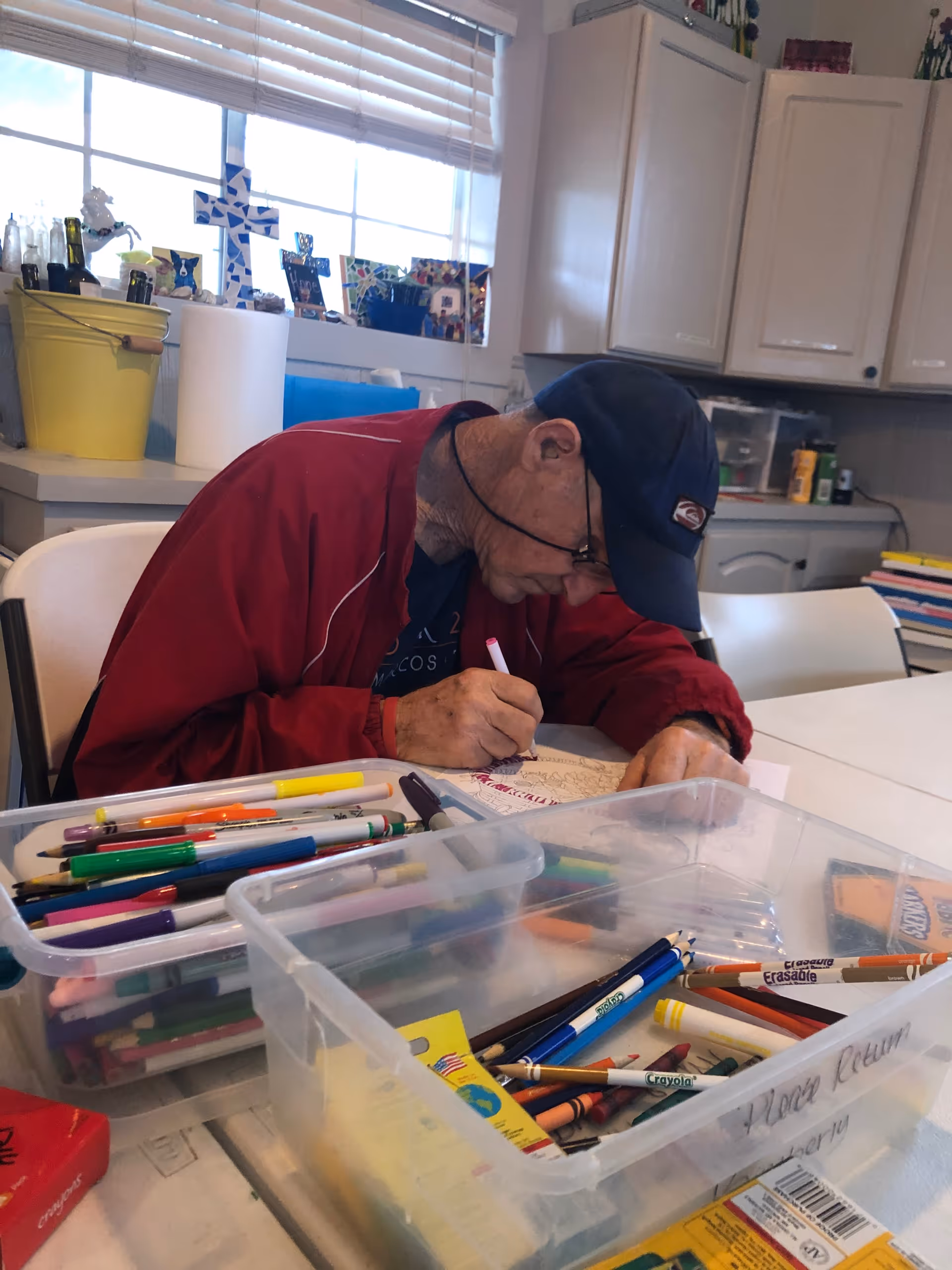 An elderly man wearing a red jacket and a navy blue cap is sitting at a table in a well-lit room, focused on coloring a detailed design with a pink marker. The table is covered with various art supplies including colored pencils, markers, and crayons organized in clear plastic containers. The background shows white cabinets, a window with blinds, and decorative items on the windowsill.