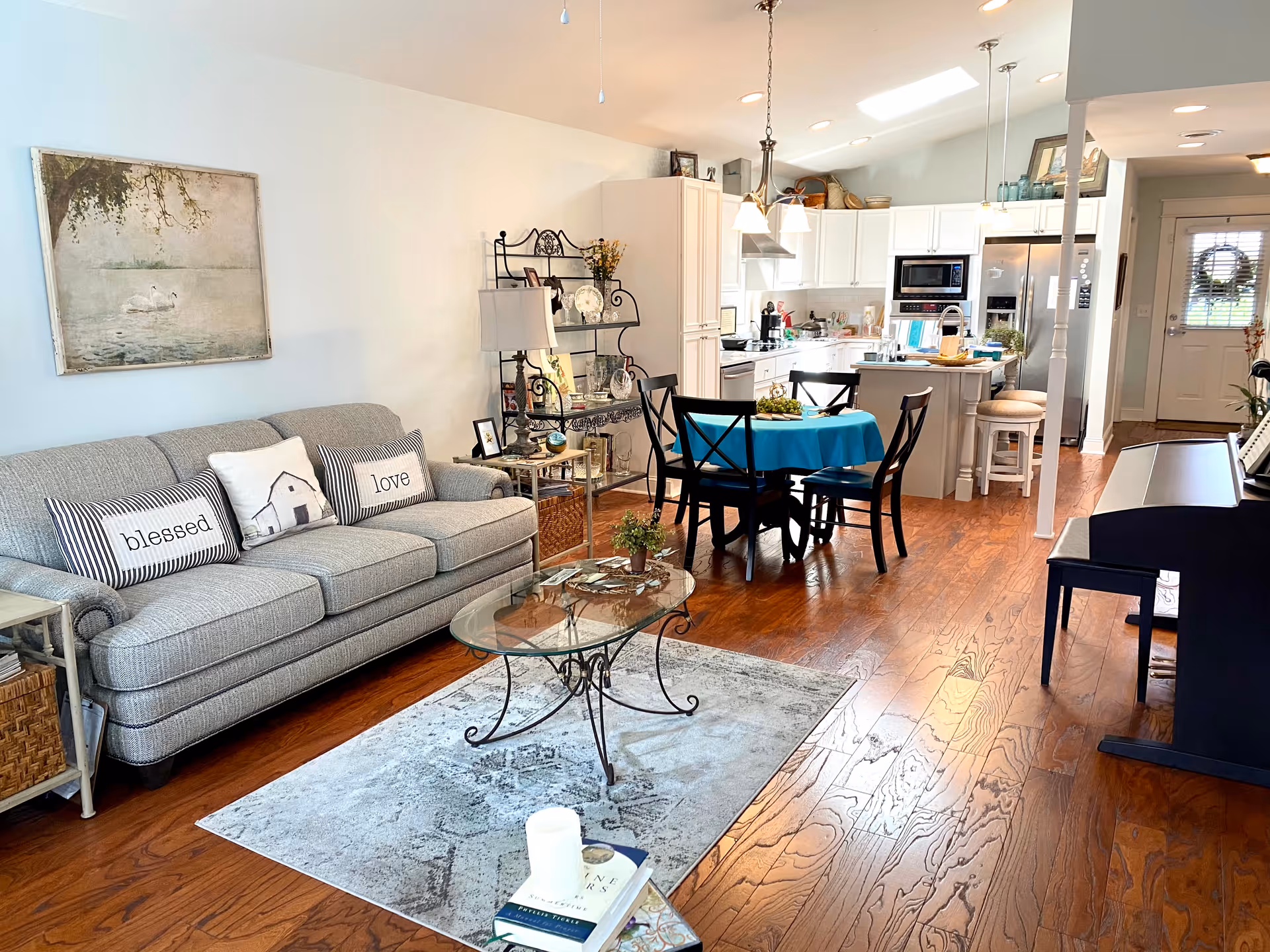 A bright and cozy open-concept living area featuring a gray sofa with decorative pillows labeled 'blessed' and 'love', a glass coffee table on a patterned rug, a black dining table with four chairs covered with a blue tablecloth, and a white kitchen with modern appliances and an island with stools. Hardwood floors and a piano are also visible, along with a front door in the background.