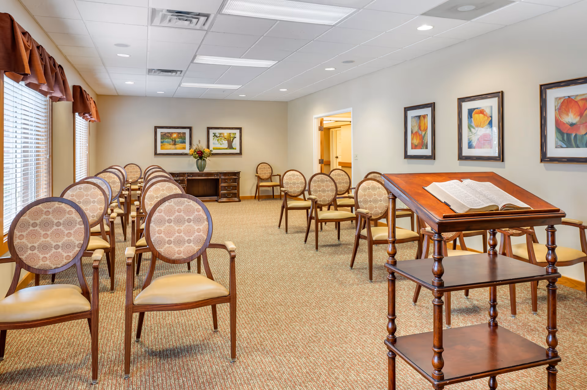 A well-lit room with rows of patterned chairs arranged facing a wooden lectern holding an open book. The room has beige walls adorned with framed floral paintings and two large windows with brown valances on the left side. A wooden sideboard with a vase of flowers is positioned against the back wall.