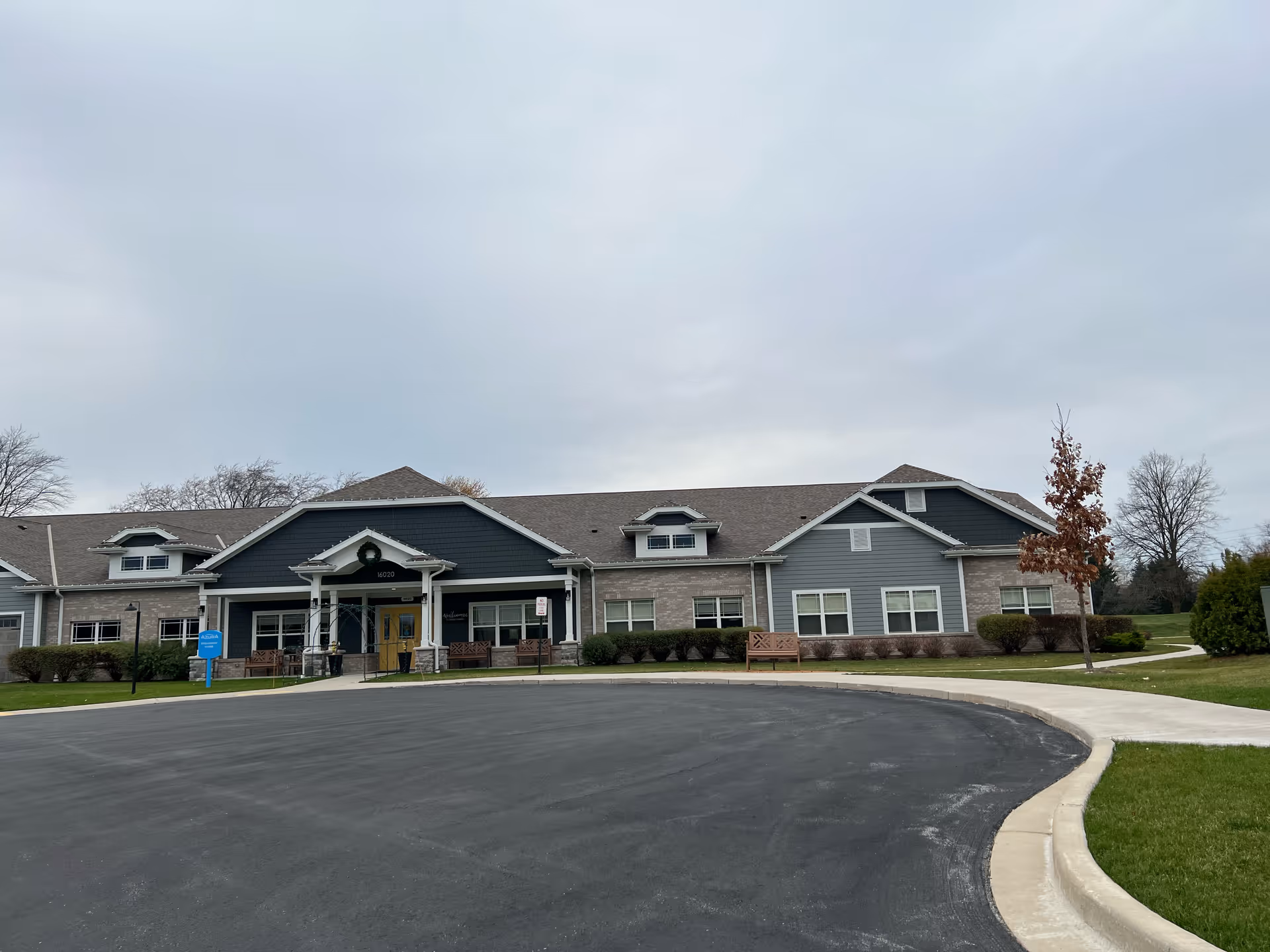 Exterior front view of a single-story assisted living facility building with a paved driveway and sidewalk leading to the entrance. The building has a combination of gray siding and brick with a peaked roof and several windows. There are benches and small bushes near the entrance, and a blue sign on the lawn that reads Azura Memory Care and Assisted Living.