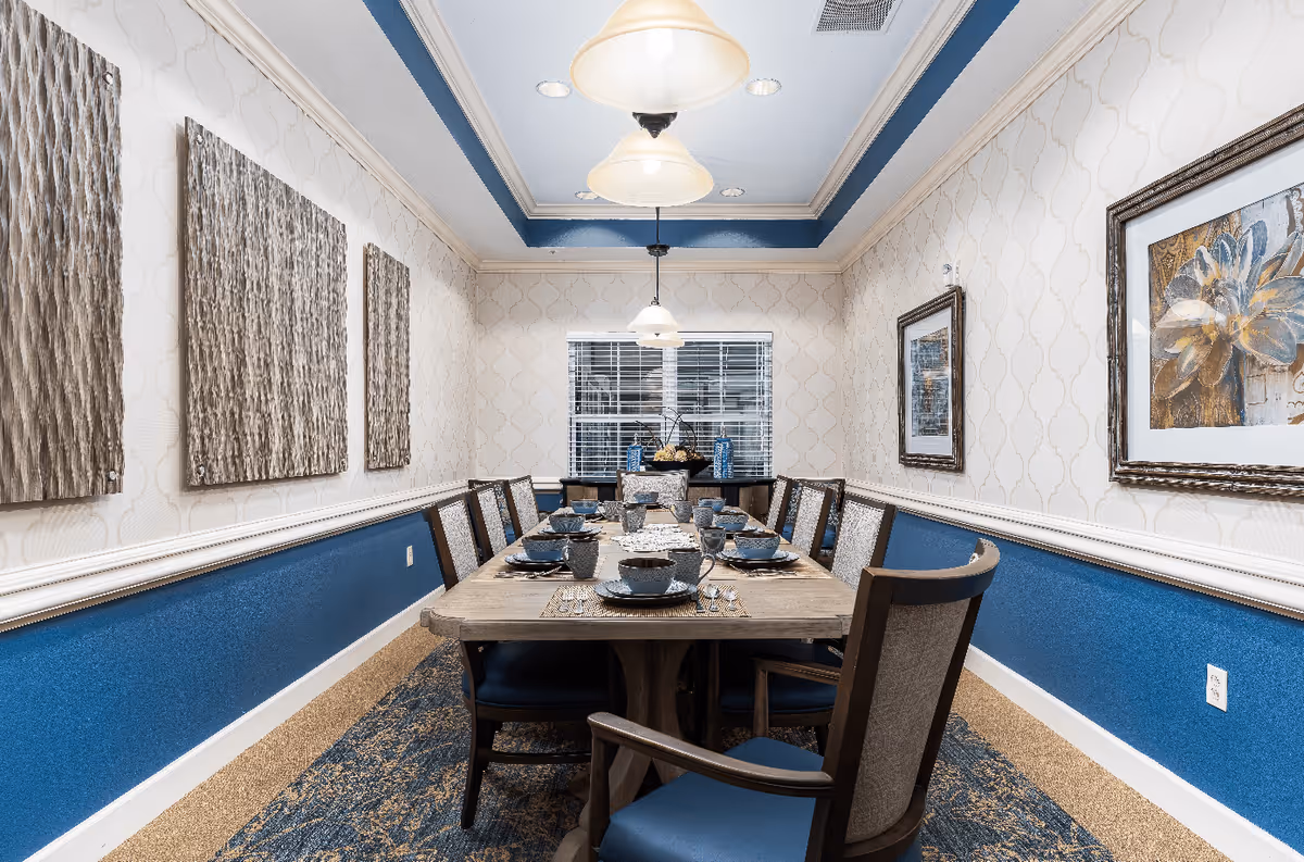 A well-lit dining room with a long wooden table set for eight people. The table is arranged with blue plates, bowls, cups, and silverware on woven placemats. The room features blue and beige walls with decorative molding, three pendant lights hanging from the ceiling, and framed artwork on the walls. A window with blinds is at the far end of the room.