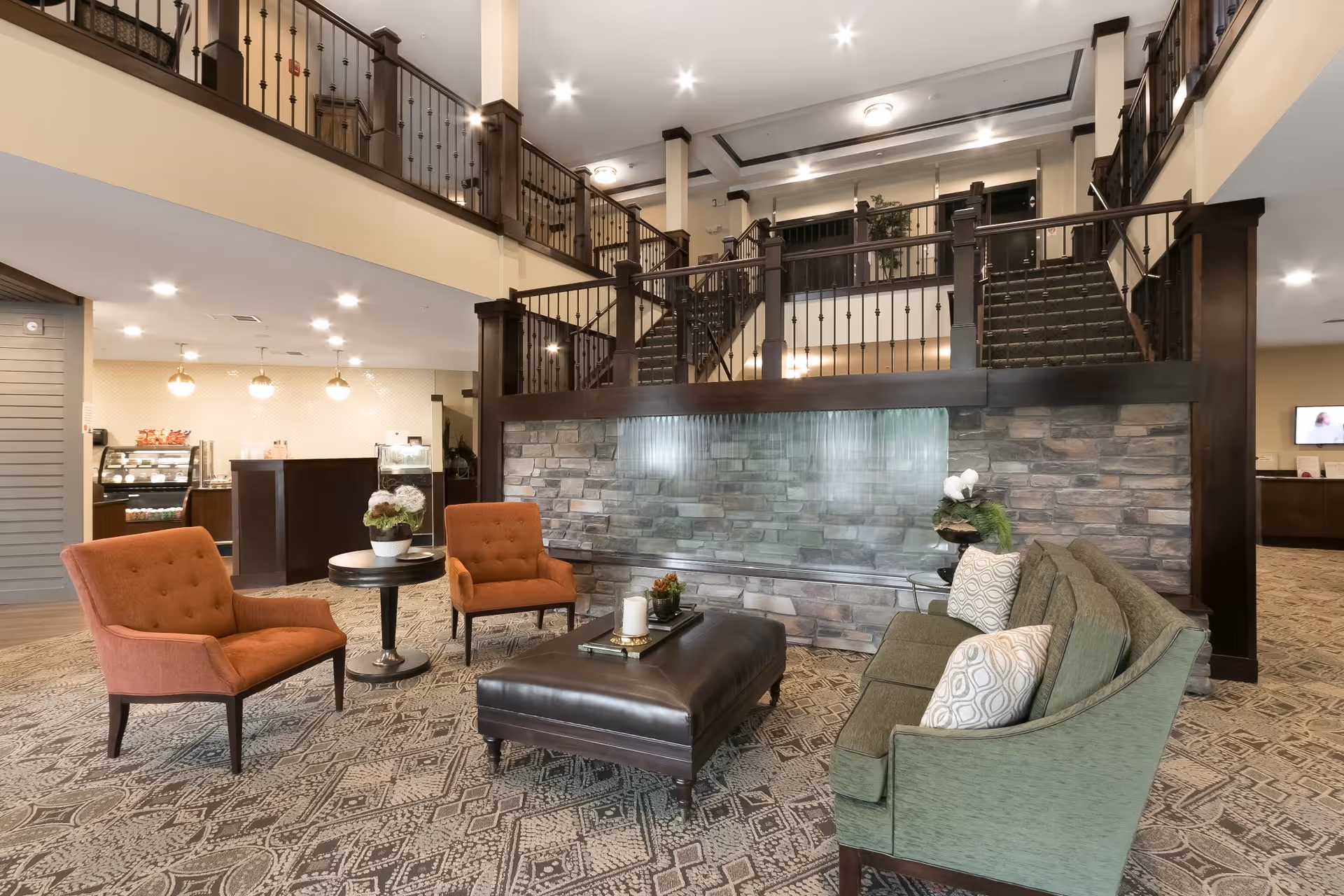 A spacious and well-lit senior living facility common area with a seating arrangement including two orange armchairs, a green sofa with patterned pillows, and a dark brown ottoman. The area features a decorative stone wall with a water feature, a patterned carpet, and a staircase with dark wooden railings leading to an upper level. In the background, there is a reception or snack area with hanging lights and display cases.