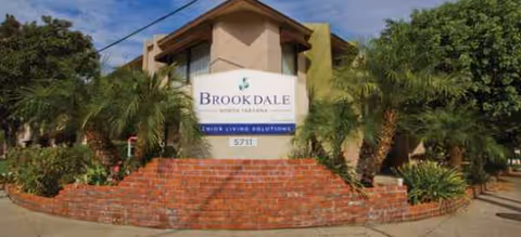 Front exterior of a senior living building with a sign and brick planter wall, palm trees.