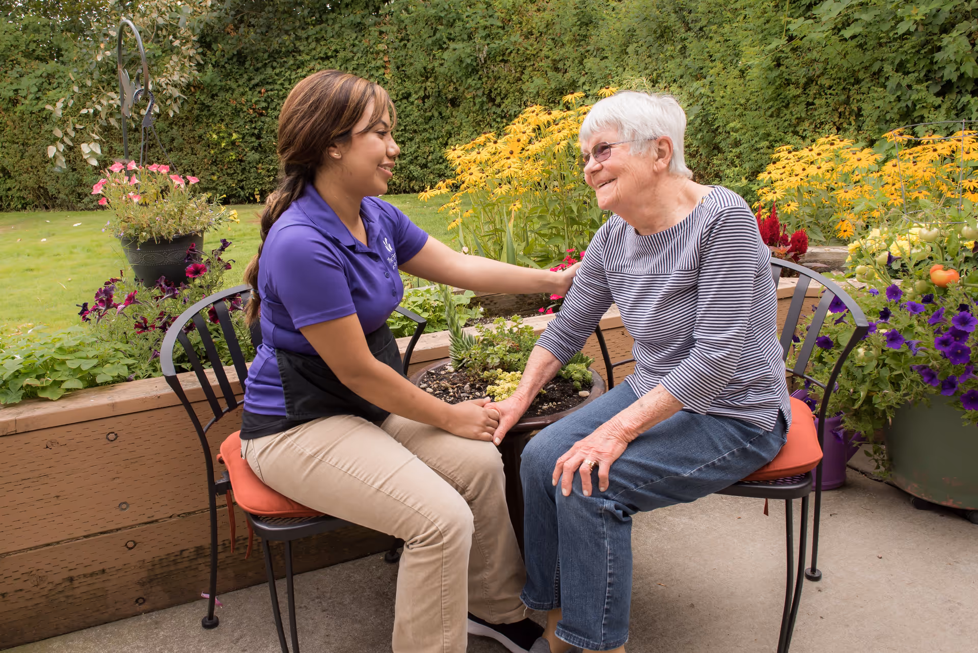 A young woman in a purple shirt and beige pants sits on a chair outdoors, holding hands and smiling warmly at an elderly woman with white hair wearing glasses, a striped shirt, and jeans. They are seated in a garden area with colorful flowers and greenery in the background.