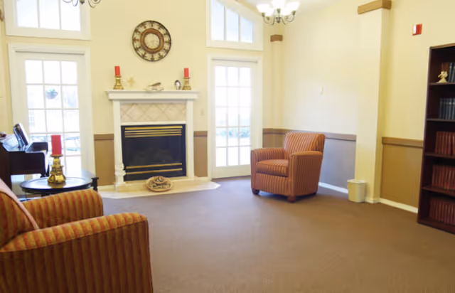 A cozy living room area with a fireplace centered on the wall, flanked by two tall windows and a glass door. Two striped armchairs are positioned facing each other on a carpeted floor. A round side table with a lamp and decorative items is next to one armchair. A bookshelf filled with books is visible on the right side of the room.