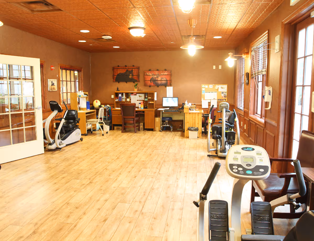 Bright multipurpose activity room with exercise machines, desks, and wooden floors in a senior living facility.