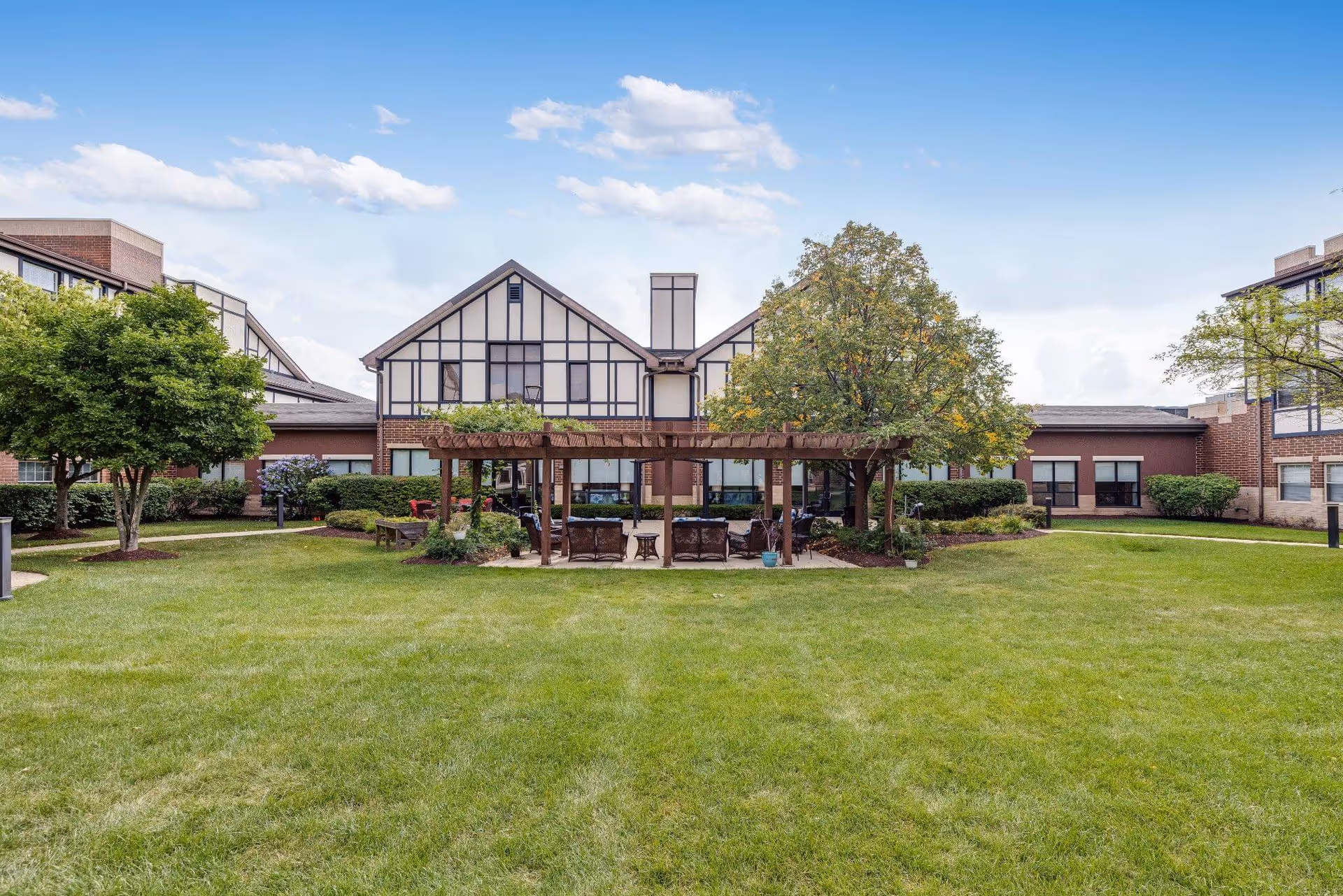 A grassy courtyard with a wooden pergola and seating flanked by trees in front of a Tudor-style senior living building.