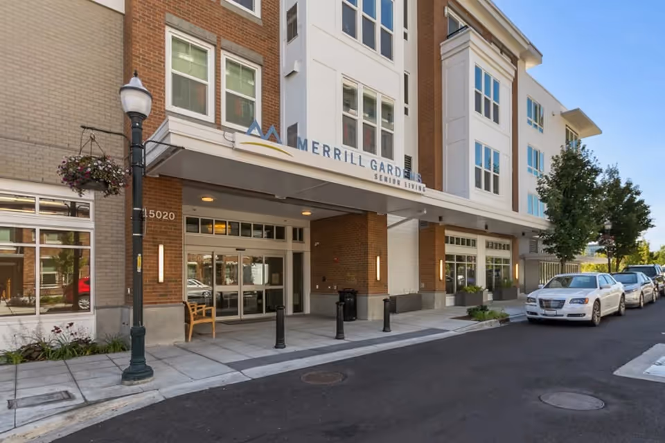 Exterior front entrance of Merrill Gardens at Burien senior living facility with a covered entryway, a bench, street lamp with hanging flower basket, and parked cars along the street.