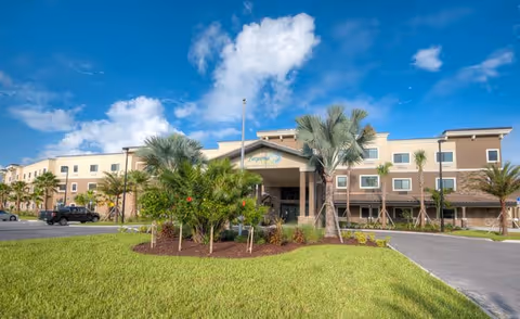 Exterior view of a multi-story senior living facility building with a covered entrance, surrounded by palm trees and landscaping under a blue sky with some clouds.