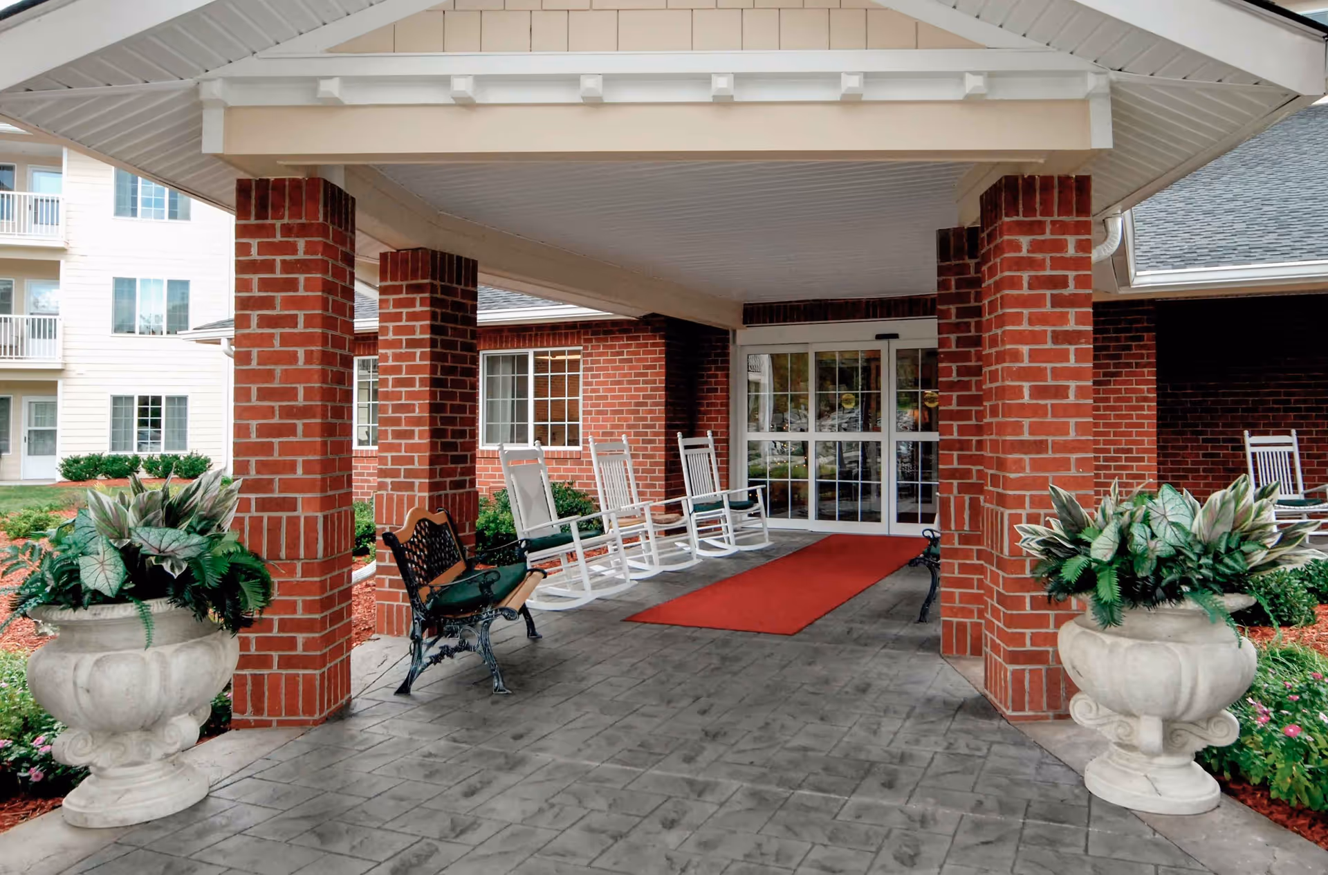 Covered entrance area of a senior living facility with brick pillars, white rocking chairs, a black metal bench, two large decorative planters with green plants, and a red carpet leading to glass double doors.