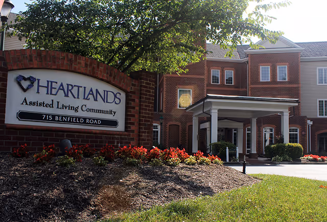 Exterior view of HeartLands Assisted Living Community building with a brick sign in the foreground displaying the facility name and address, surrounded by landscaping with flowers and a tree.