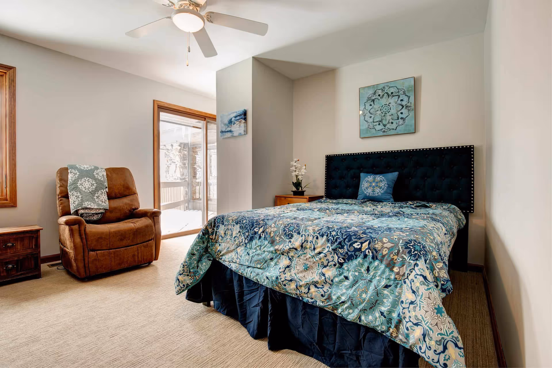 A bedroom with a large bed covered in a blue and teal patterned comforter and matching pillow. There is a dark upholstered headboard with nailhead trim. To the left of the bed is a wooden nightstand with a small plant on it. A brown recliner chair with a patterned throw blanket is positioned near a window and sliding glass door that leads outside. The walls are light-colored, and there is a ceiling fan with a light fixture above.