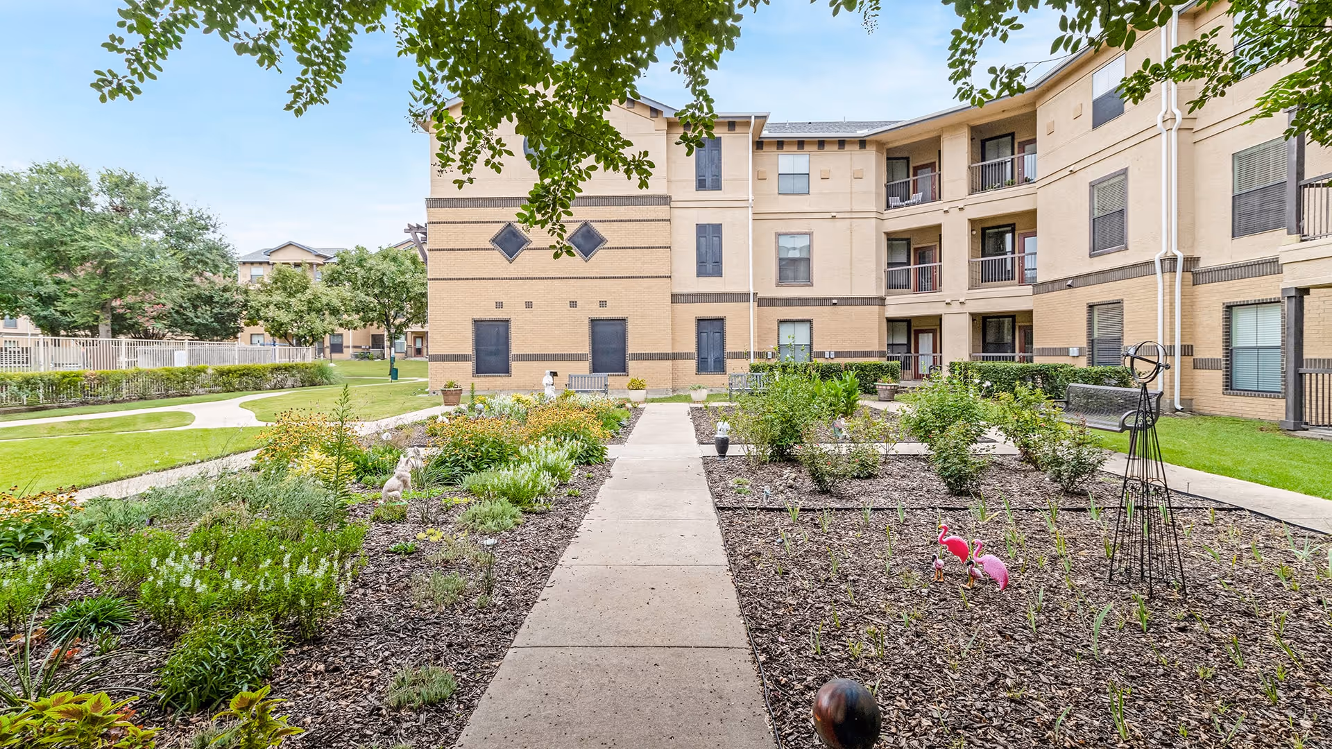 Outdoor garden area with a concrete walkway leading through planted flower beds and shrubs, surrounded by a three-story beige apartment building with balconies and windows. There are decorative garden ornaments including pink flamingos and a metal sculpture, with green grass and trees in the background under a clear sky.