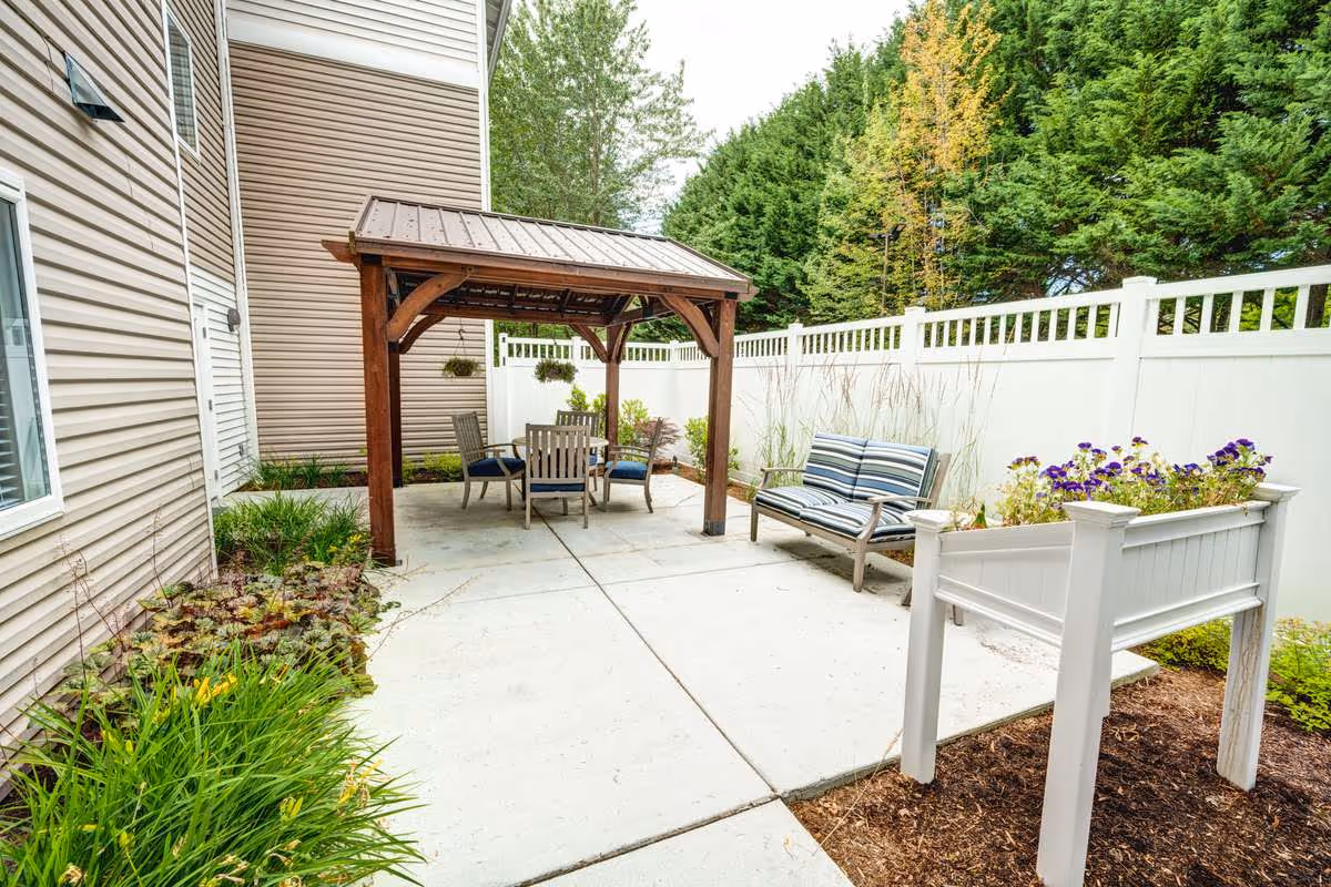 Outdoor patio area at The Gardens at Marysville featuring a wooden pergola with a table and four chairs underneath, a cushioned bench with striped upholstery, white privacy fencing, and planters with flowers and greenery.