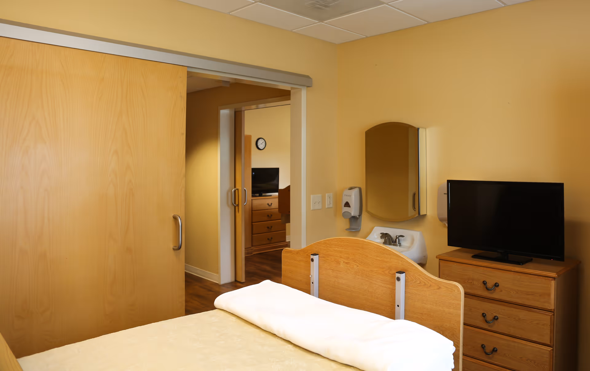 A simple patient bedroom with a wooden bed, dresser and TV, wall-mounted sink and mirror, and a sliding wooden door.