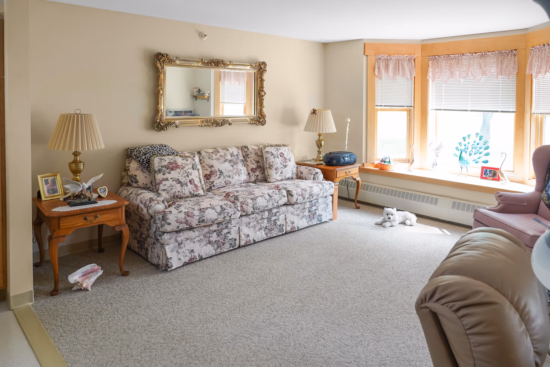 Sunlit living room with a floral-patterned sofa flanked by wooden side tables and lamps, an ornate mirror above, a bay window seat with decorations, and a stuffed toy on the carpet.