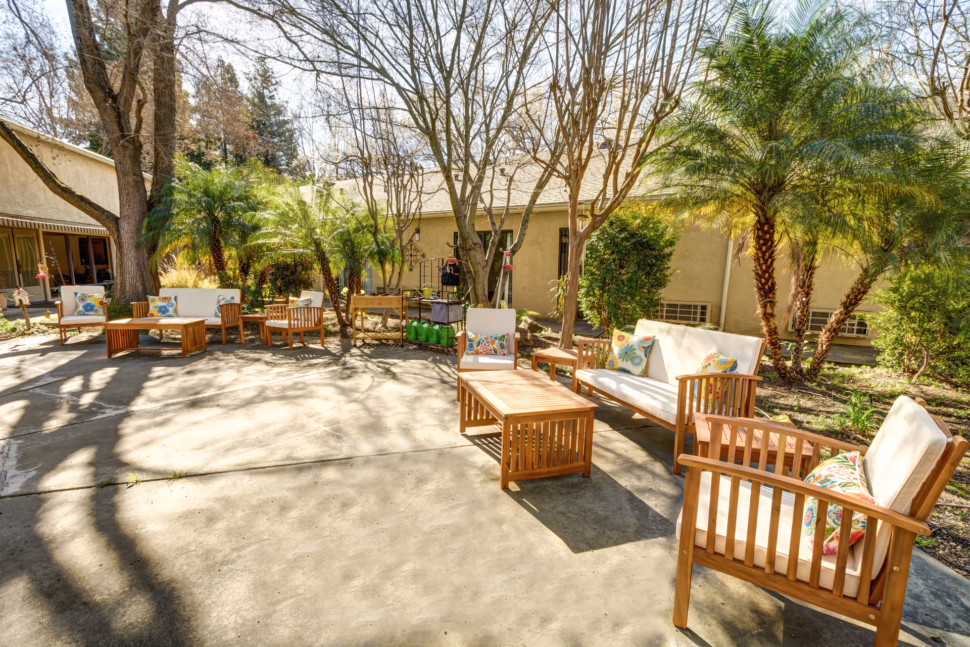 Outdoor patio area at Greenhaven Estates Assisted Living and Memory Care featuring wooden outdoor furniture with white cushions and colorful floral pillows arranged around wooden coffee tables. The patio is surrounded by trees, palm plants, and bushes with a beige building in the background under a clear sky.