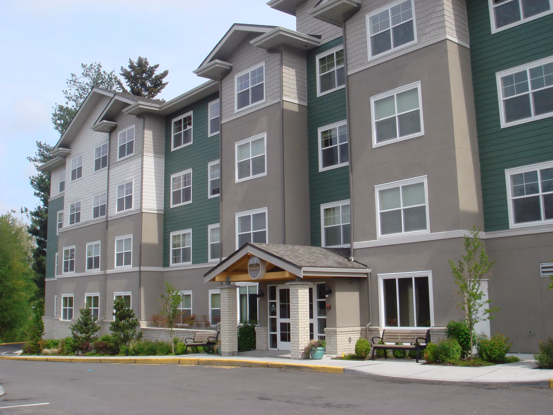 Exterior view of a multi-story senior living facility named The Creekside, featuring a combination of green and beige siding, multiple windows, a covered entrance with a sign, benches, and landscaped greenery around the building.